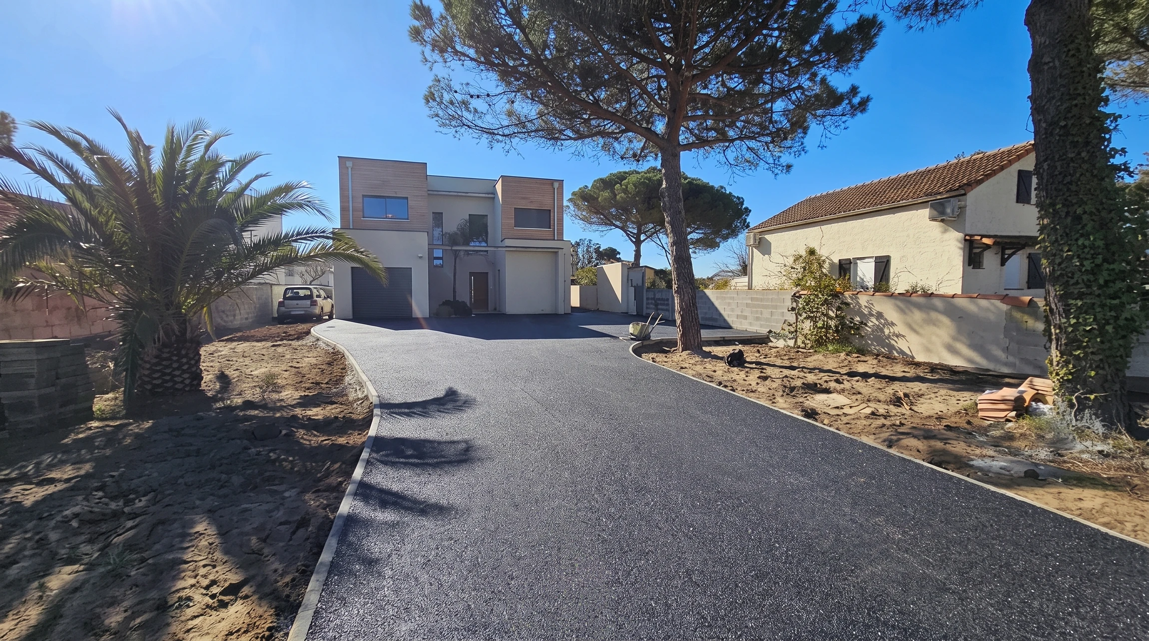 Allée privative neuve en enrobé bitumineux noir, bordurettes blanches, menant à une villa contemporaine à bardage bois, double garage