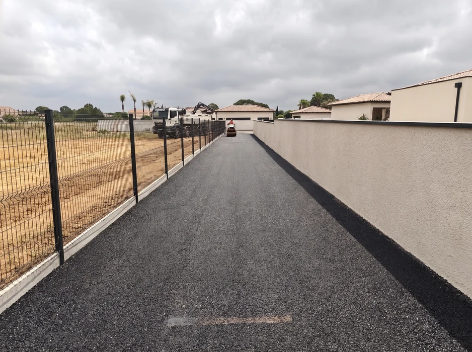 Pose d'enrobé bitumineux sur une allée privée entre un mur de clôture en enduit et une clôture rigide à panneaux soudés gris anthracite sur platines