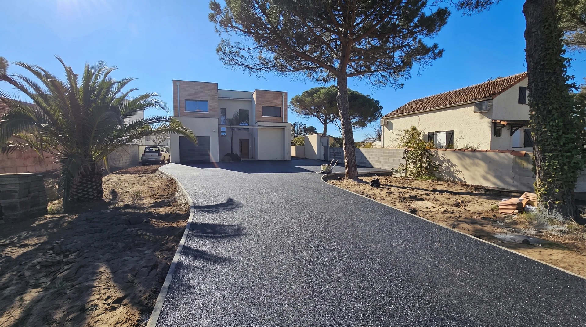 Allée privative neuve en enrobé bitumineux noir, bordurettes blanches, menant à une villa contemporaine à bardage bois, double garage