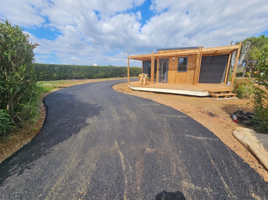 Allée privée en enrobé bitumineux noir fraîchement posé, tracé courbe desservant un chalet en bois moderne avec terrasse en bardage pin naturel et structure poteau-poutre