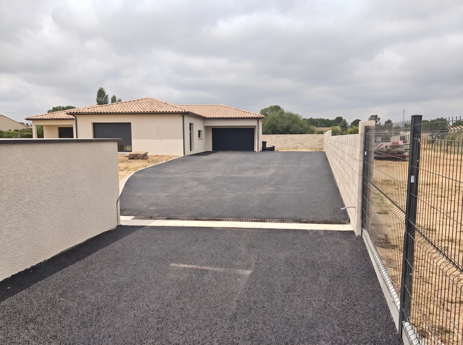 Allée de garage en enrobé bitumineux noir fraîchement posé, devant une maison plain-pied avec toiture en tuiles canal
