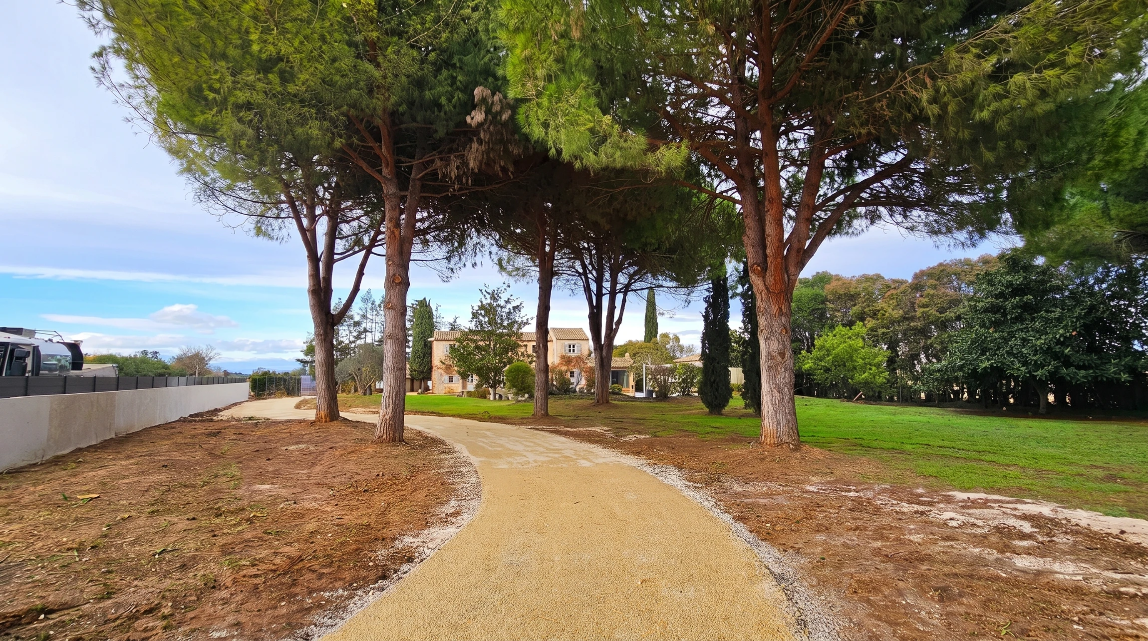 Allée d'accès courbe en stabilisé beige, bordée de caniveaux en graviers blancs, traversant une pinède de pins parasols aux troncs rougeâtres