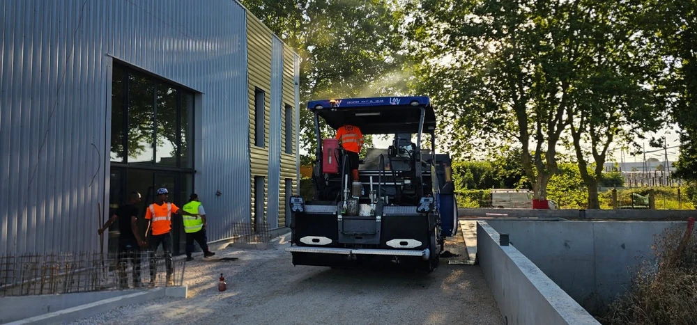 Chantier extérieur de voirie, finisseur à asphalte bleu de marque LAV en action entre un bâtiment industriel à bardage métallique et un mur en béton
